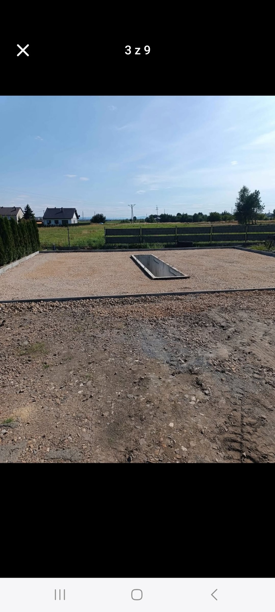 Przygotowany teren pod budowę: wyrównany, utwardzony żwirem, obrzeża z kostki, z widocznym betonowym elementem w ziemi. W tle domy i zieleń.