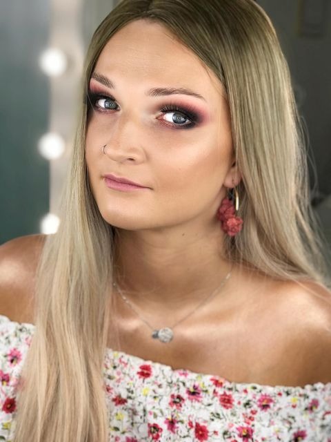 Portret kobiety z intensywnym makijażem oczu w odcieniach różu i czerni, subtelnym makijażem ust, blond włosami, kolczykami z pomponami i srebrnym naszyjnikiem na tle rozmytego lustra.