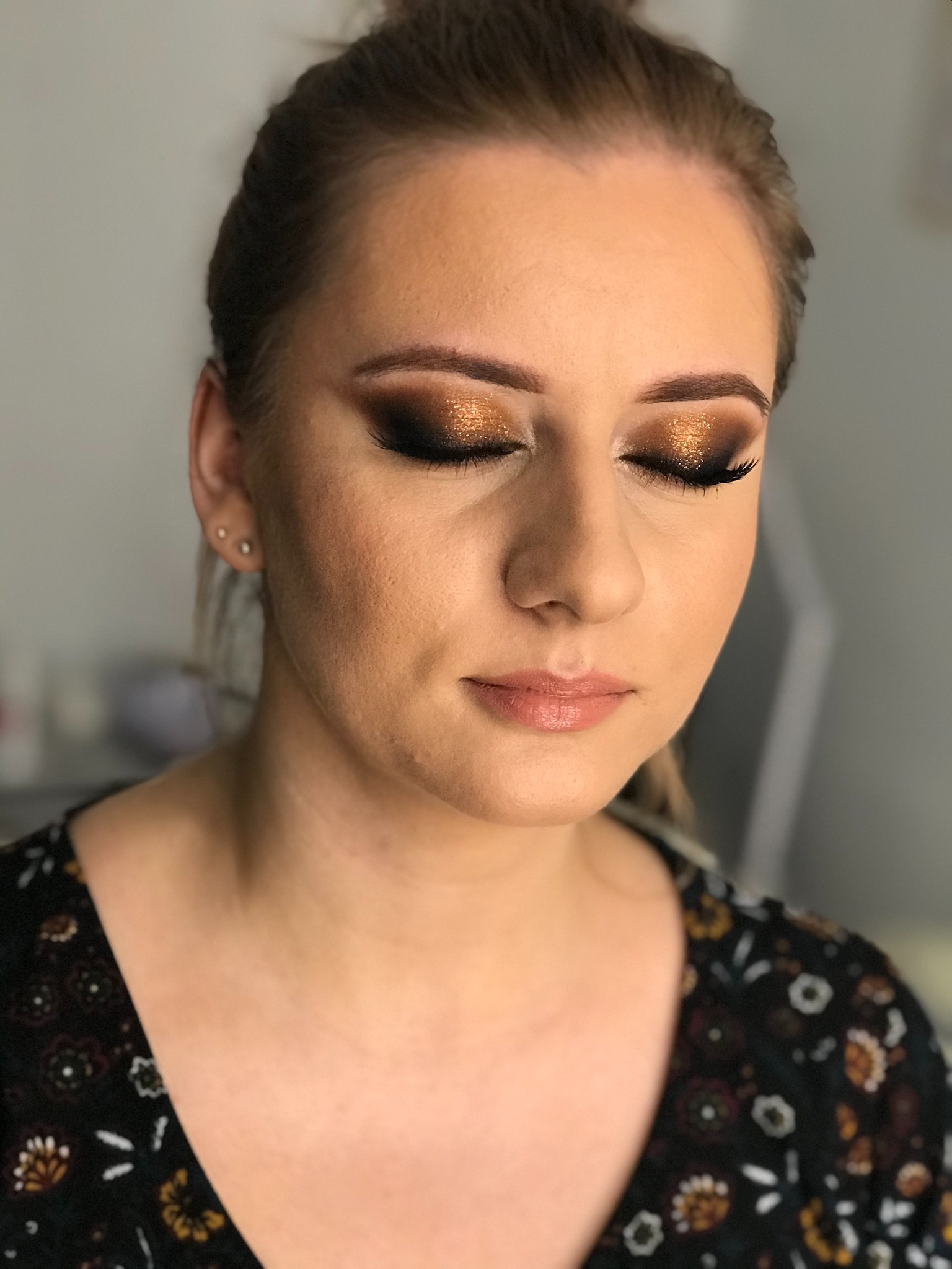 Kobieta z zamkniętymi oczami prezentuje makijaż oczu w odcieniach brązu i złota z efektem brokatu, podkreślony czarnym eyelinerem i sztucznymi rzęsami; delikatny makijaż ust w odcieniu nude.