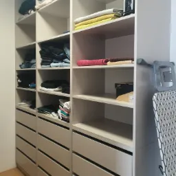 Wysoka, jasnoszara szafa z otwartymi półkami i szufladami, wypełniona ubraniami, ręcznikami, żelazkiem i deską do prasowania.