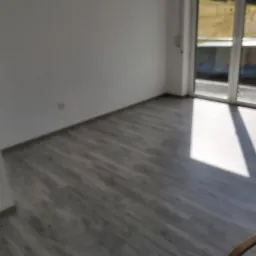 Jasne wnętrze z szarymi panelami podłogowymi, widoczne białe ściany i okno balkonowe z widokiem na zieleń, silne światło słoneczne.