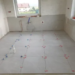 Szare płytki podłogowe w trakcie układania, z widocznymi krzyżykami dystansowymi i butelką środka czyszczącego w rogu pomieszczenia, ściany częściowo wykończone płytkami, okno z widokiem na dom.