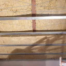 Poddasze z wełną mineralną, zabezpieczoną sznurkiem i metalowym stelażem, widoczne elementy drewniane konstrukcji.