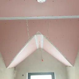 Montaż różowej płyty gipsowo-kartonowej na skośnym poddaszu z widocznymi profilami narożnymi i otworami po wkrętach, surowy stan wnętrza.