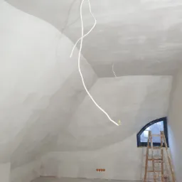 Poddasze po nałożeniu gładzi, z widocznymi przewodami elektrycznymi zwisającymi z sufitu, drewnianą drabiną opartą o okno w kształcie łuku i gniazdkami elektrycznymi w ścianie.