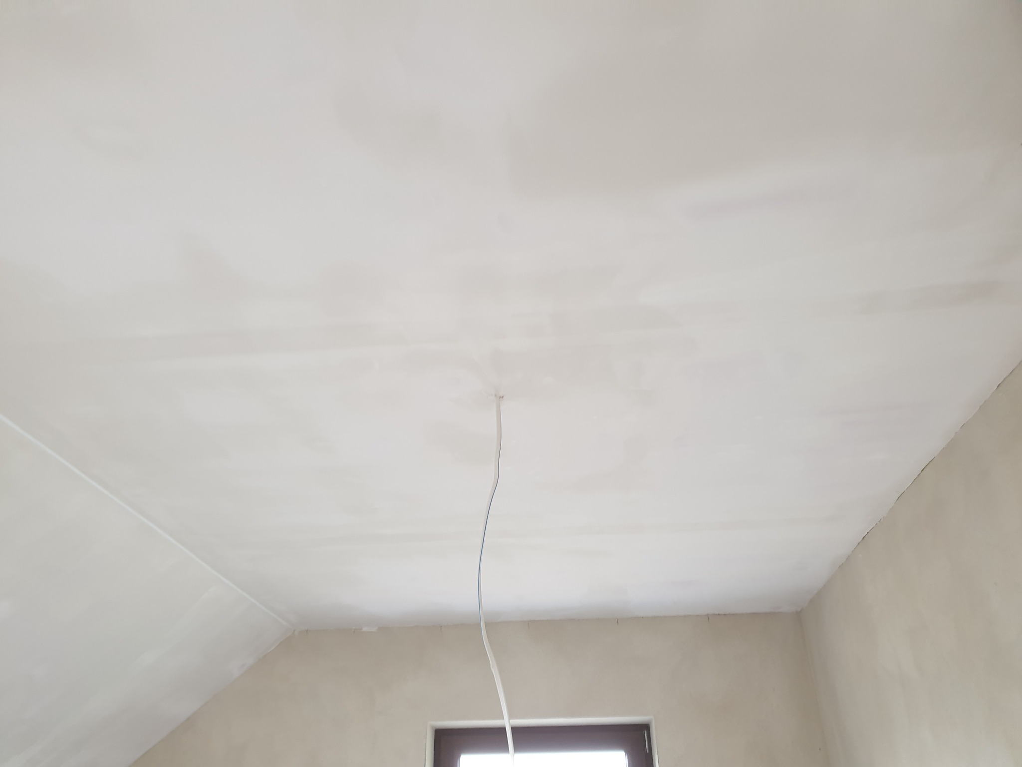 Gładka, biała powierzchnia sufitu po nałożeniu gładzi, widoczny wystający kabel elektryczny i fragment okna w dolnej części kadru.