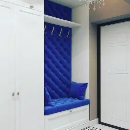 Narożnik w przedpokoju z niebieską, pikowaną tapicerką na ścianie i siedzisku, uzupełniony o wieszaki i półkę, z widocznym fragmentem białej szafy i drzwi.