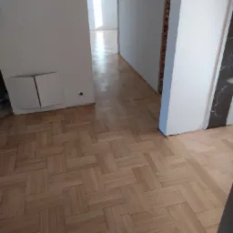 Ułożony parkiet w jodełkę francuską w jasnym odcieniu, widoczne niedokończone ściany i przygotowania do wykończenia wnętrza.