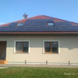 Dom jednorodzinny z panelami fotowoltaicznymi na czerwonym dachu, widoczne dwa okna dachowe oraz antena satelitarna, trawnik przed domem.