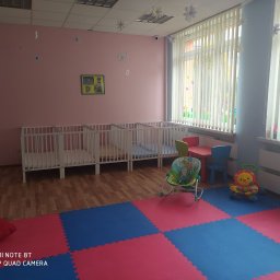 AKADEMIA MALUCHA DANUTA SKOMOROWSKA-LEŹNICKA - Wnętrze pokoju dziecięcego z kilkoma białymi łóżeczkami, kolorową podłogą z puzzli piankowych, krzesełkiem do karmienia i zabawkami, dekoracjami kwiatowymi na ścianach i suficie, oraz pionowymi...