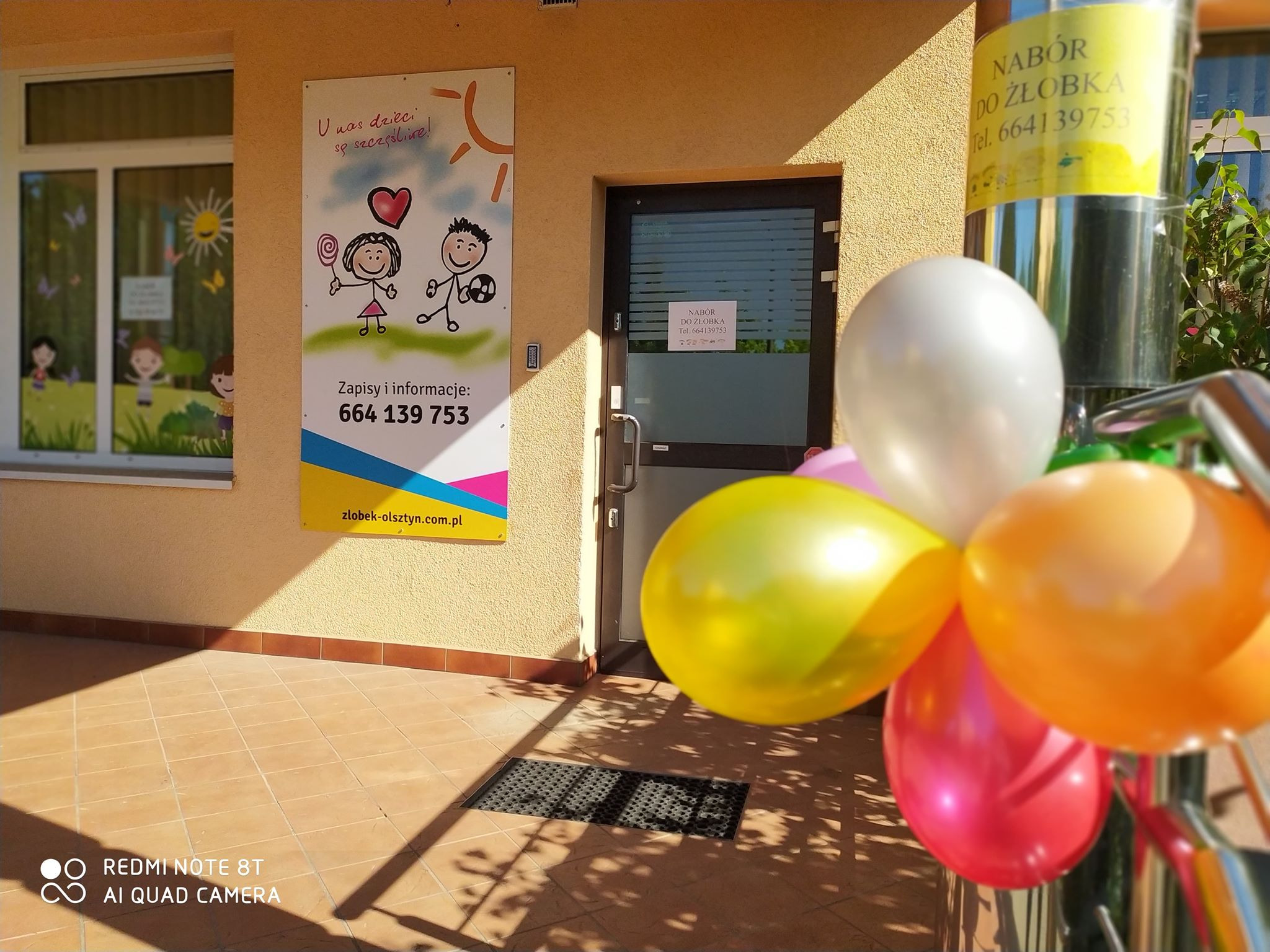 Wejście do placówki z kolorowymi balonami, plakat z informacją o naborze i rysunkami dzieci oraz numerem telefonu.