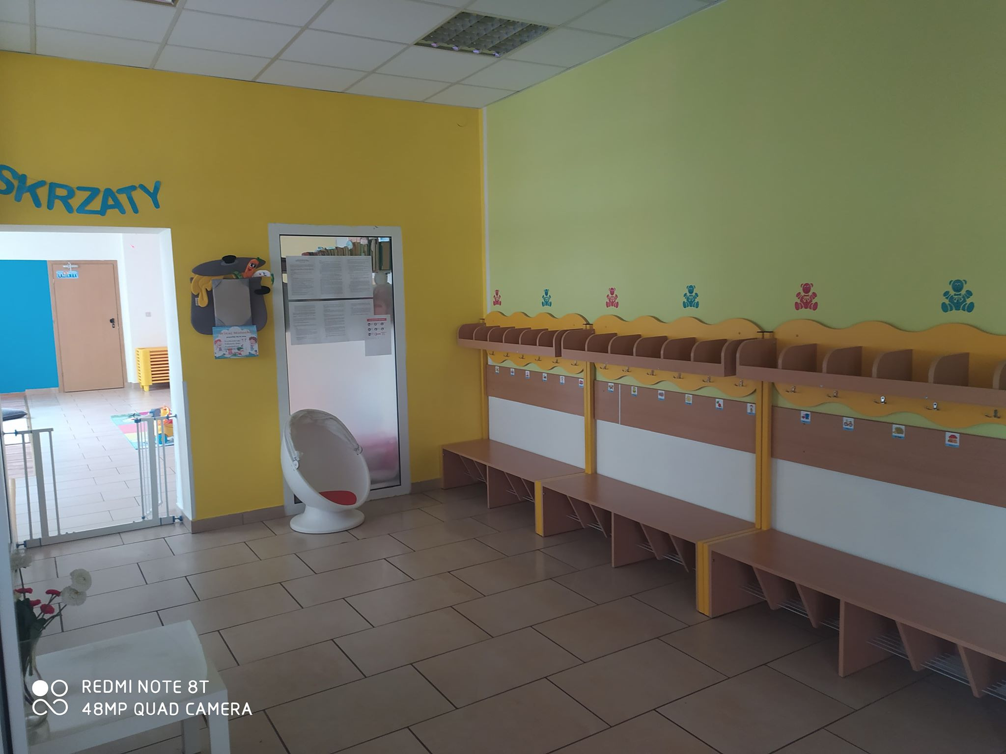 Szatnia w przedszkolu z żółtymi i zielonymi ścianami, wieszakami z motywem misia i miejscami do siedzenia, z napisem 'Skrzaty' nad wejściem.