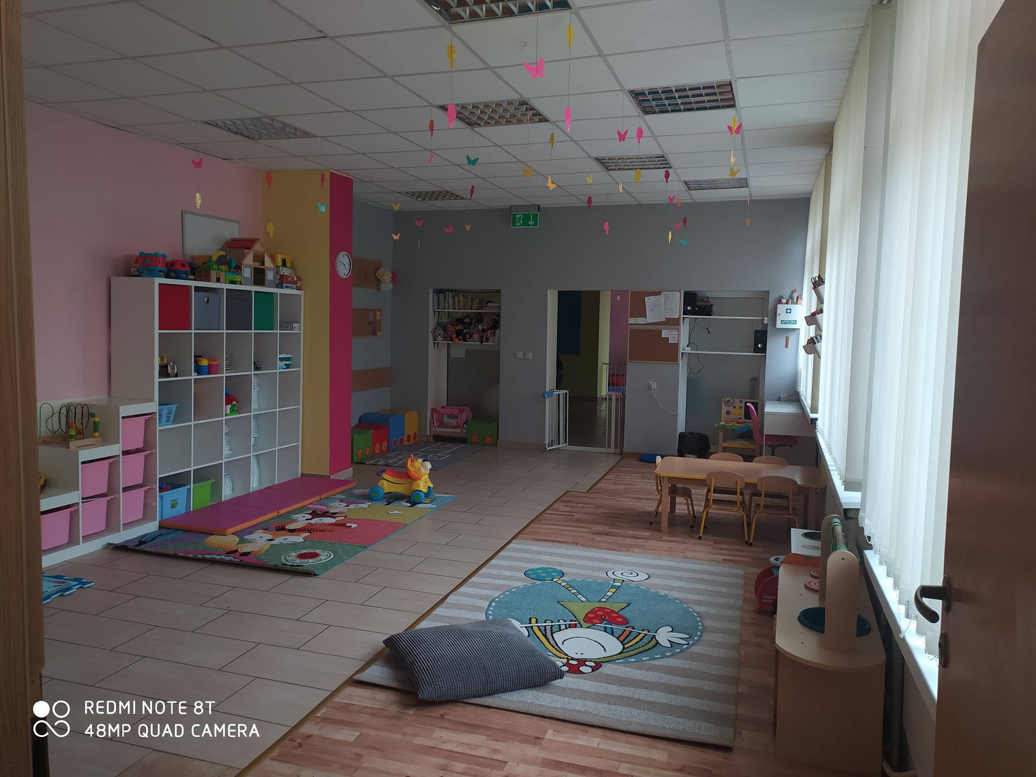 Wnętrze sali zabaw z kolorowymi motylami zawieszonymi pod sufitem, regałami z zabawkami, dywanami z postaciami z bajek i stolikami dla dzieci.