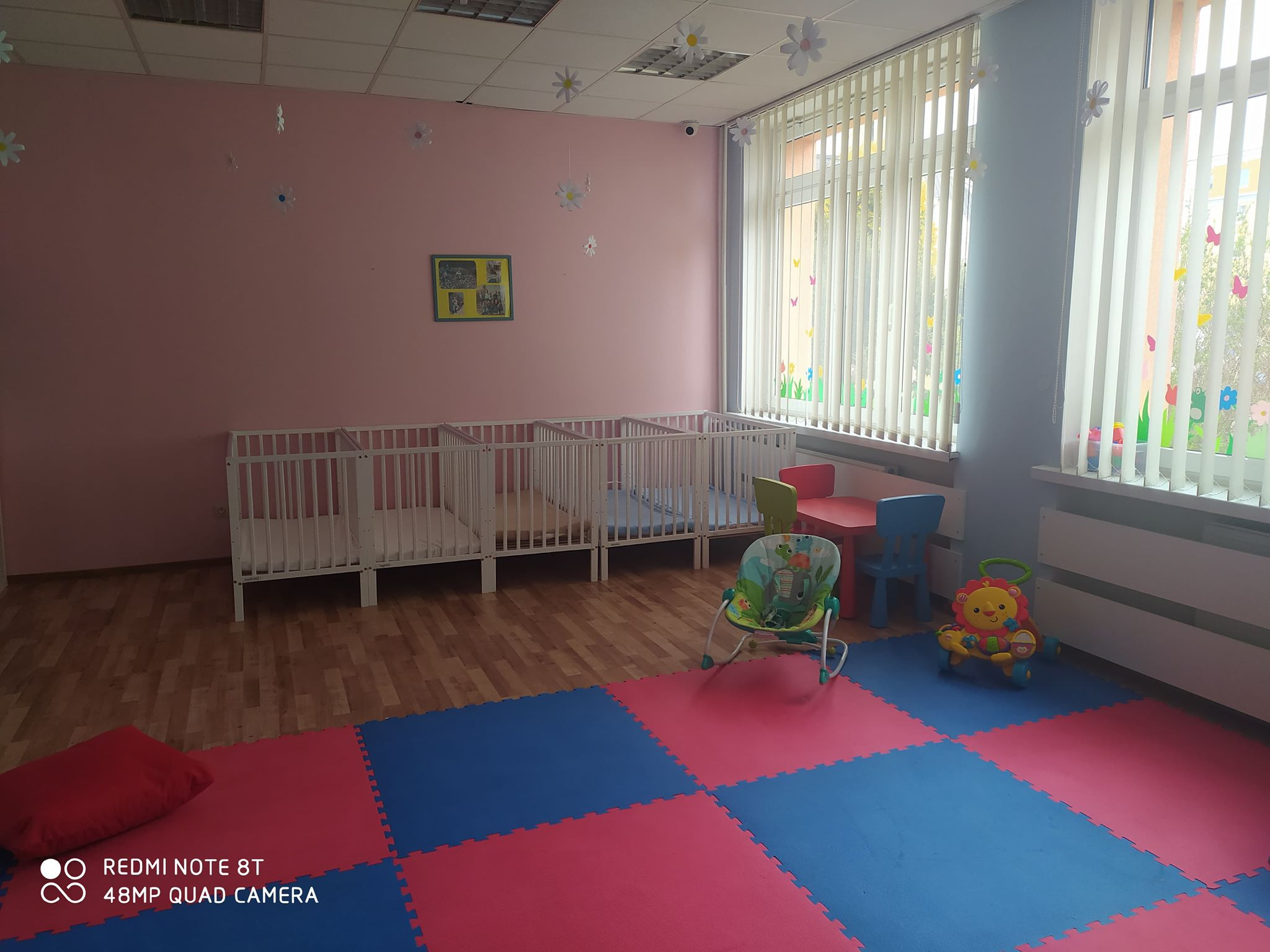 Wnętrze pokoju dziecięcego z kilkoma białymi łóżeczkami, kolorową podłogą z puzzli piankowych, krzesełkiem do karmienia i zabawkami, dekoracjami kwiatowymi na ścianach i suficie, oraz pionowymi...