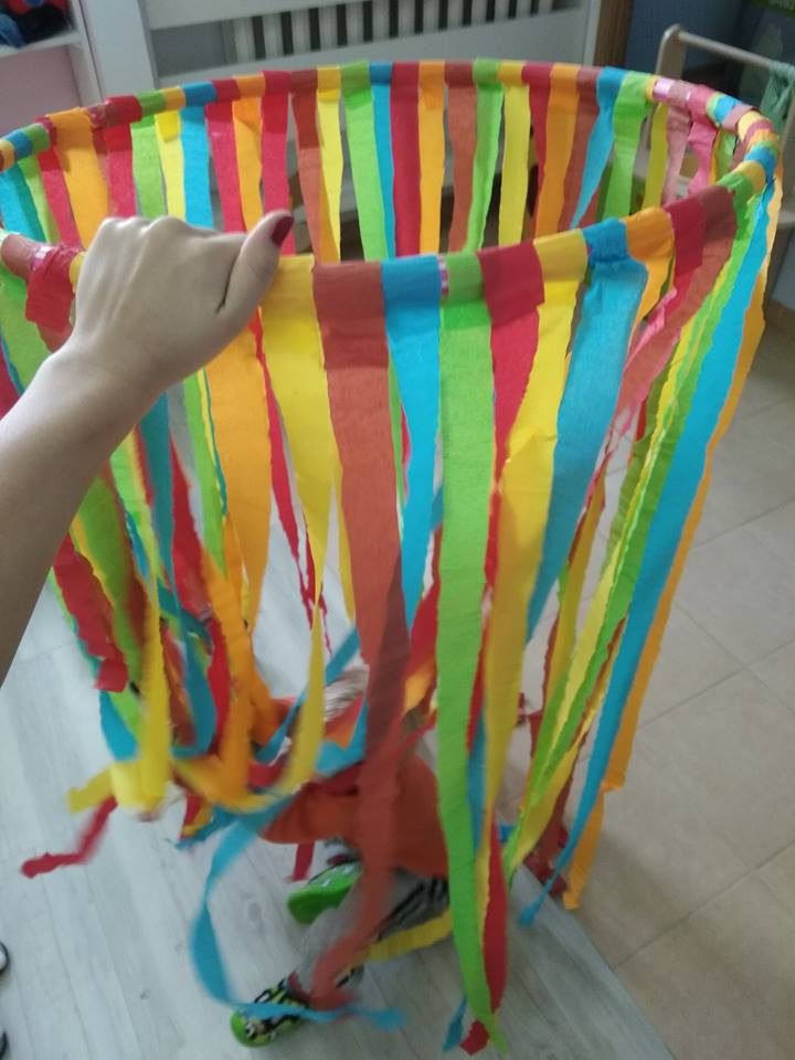 Ręka trzyma obręcz ozdobioną kolorowymi, papierowymi frędzlami, pod którą widać fragment dziecka bawiącego się na podłodze.