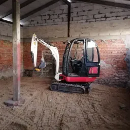 Mini koparka JCB wewnątrz budynku z ceglanymi ścianami i ziemią na podłodze, widoczne ślady gąsienic na piasku, oświetlenie naturalne z okna po prawej.