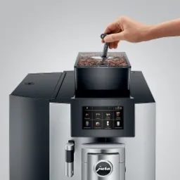 Automatyczny ekspres do kawy Jura z pojemnikiem na ziarna kawy i dłonią regulującą ustawienia, widoczny panel sterowania z ikonami różnych rodzajów kaw.