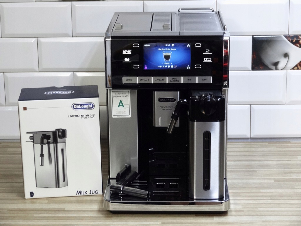 Automatyczny ekspres do kawy Delonghi Primadonna Exclusive z pojemnikiem na mleko i pudełkiem LatteCrema System na drewnianym blacie na tle białych płytek.