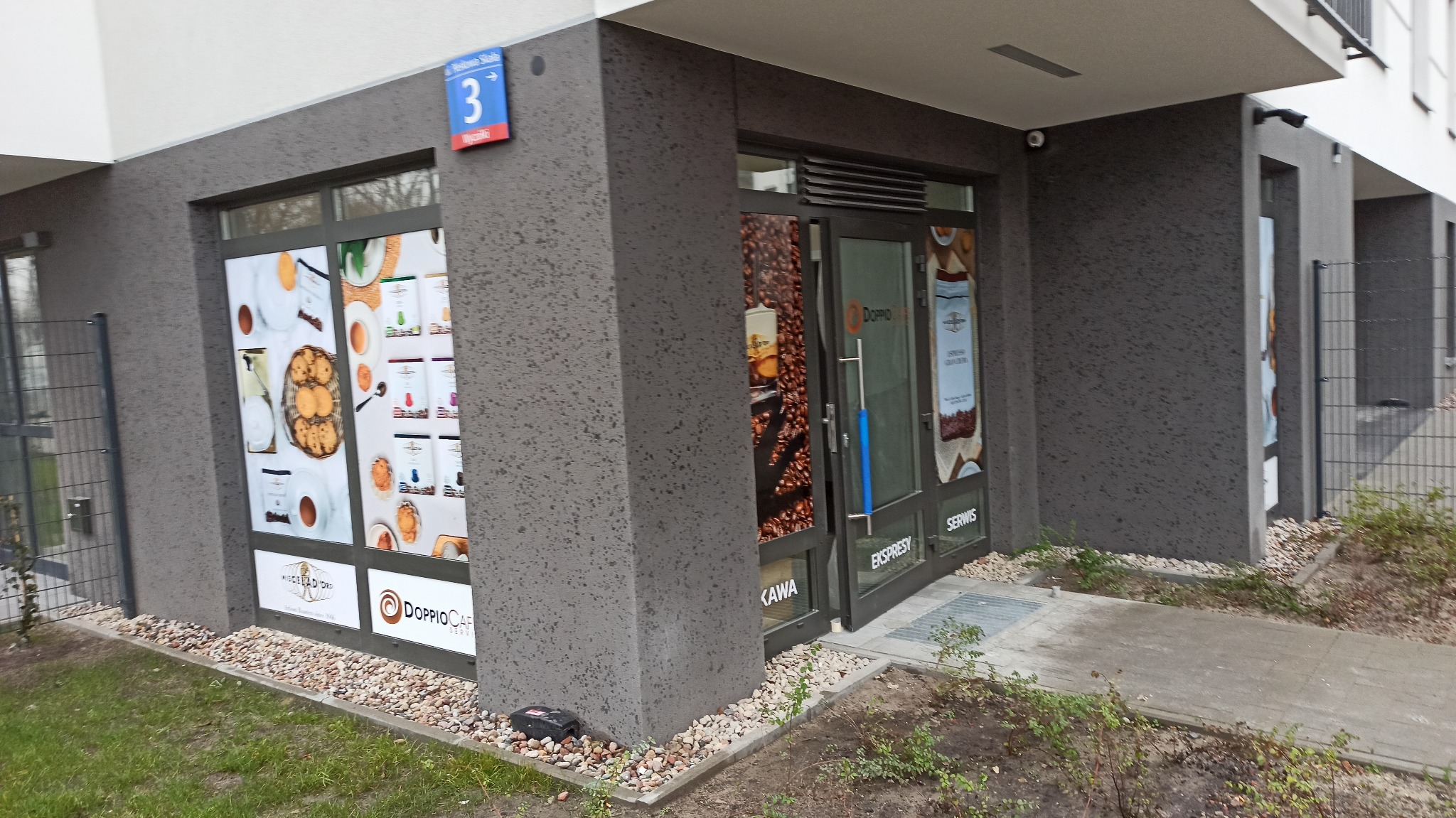 Fasada budynku z napisem 'Serwis Ekspresów' na drzwiach wejściowych, oklejone witryny z grafikami kawy i ciasteczek, widoczny adres budynku.