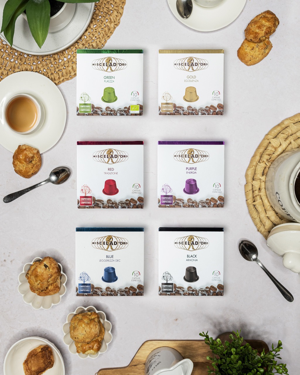 Kompozycja z sześcioma opakowaniami kapsułek do kawy Miscelad'Oro w różnych kolorach (zielony, złoty, czerwony, fioletowy, niebieski, czarny), filiżanka espresso, ciasteczka i srebrne łyżeczki...