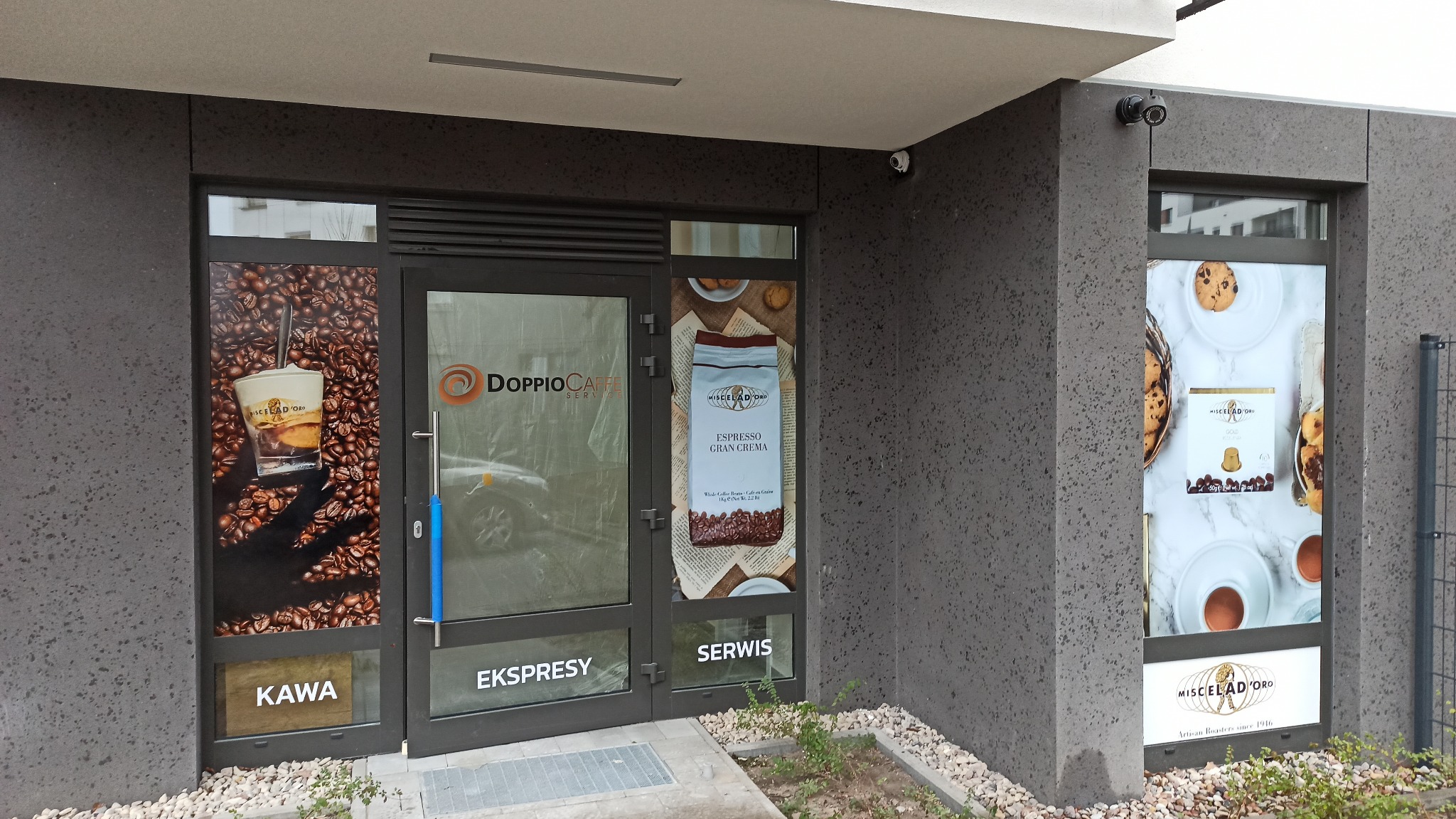 Fasada lokalu z naklejkami reklamowymi kawy, ekspresów i serwisu ekspresów do kawy, widoczna nazwa 'DOPPIOCAFE SERWIS', 'MISCELAD'ORO Artisan Roasters since 1916', 'KAWA', 'EKSPRESY', 'SERWIS'...