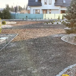 Przygotowanie terenu pod trawnik: wyrównana ziemia, obrzeża z kamieni, nasadzenia iglaków i krzewów ozdobnych w ogrodzie przydomowym.