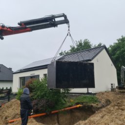 HKS- INFRASTRUKTURA Sławomir Hebda - Ciężki, czarny zbiornik zawieszony na łańcuchach dźwigu, opuszczany do wykopu obok domu z panelami słonecznymi na dachu, w tle koparka Volvo, widoczni pracownicy w ubraniach roboczych.