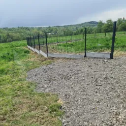 Nowo postawione ogrodzenie panelowe z podmurówką na nierównym terenie, widok z drogi szutrowej w kierunku zalesionych wzgórz pod pochmurnym niebem.