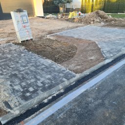 HKS- INFRASTRUKTURA Sławomir Hebda - Fragment układanej nawierzchni z szarej kostki brukowej, widoczny piasek i podsypka, z boku skrzynka elektryczna zabezpieczona taśmą, w tle hałda ziemi i częściowo ukończony budynek.
