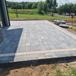 HKS- INFRASTRUKTURA Sławomir Hebda - Nowoczesny taras z szarej kostki brukowej, obrzeża z jasnych kostek. Widok na trawnik i drzewa. Częściowo ułożona kostka, paleta z kostką w tle.