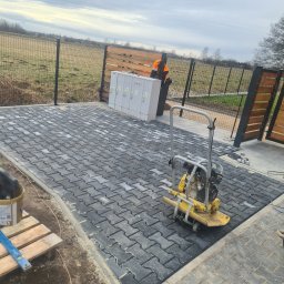HKS- INFRASTRUKTURA Sławomir Hebda - Ułożona, szara kostka brukowa na posesji z ogrodzeniem. Widoczna zagęszczarka, skrzynka elektryczna i częściowo osoba w odblaskowej kamizelce. Pochmurne niebo w tle.