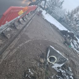 HKS- INFRASTRUKTURA Sławomir Hebda - Zimowy krajobraz z wykopem pod kanalizację deszczową, widoczne betonowe studzienki, gruz i maszyna budowlana w tle. Prace ziemne w toku, zabezpieczenia na drodze.