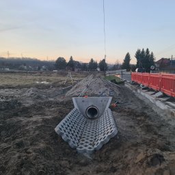 HKS- INFRASTRUKTURA Sławomir Hebda - Betonowy przepust rurowy z ażurowym korytem w wykopie, obok droga z barierami. W tle teren budowy, drzewa i zabudowania pod wieczornym niebem.
