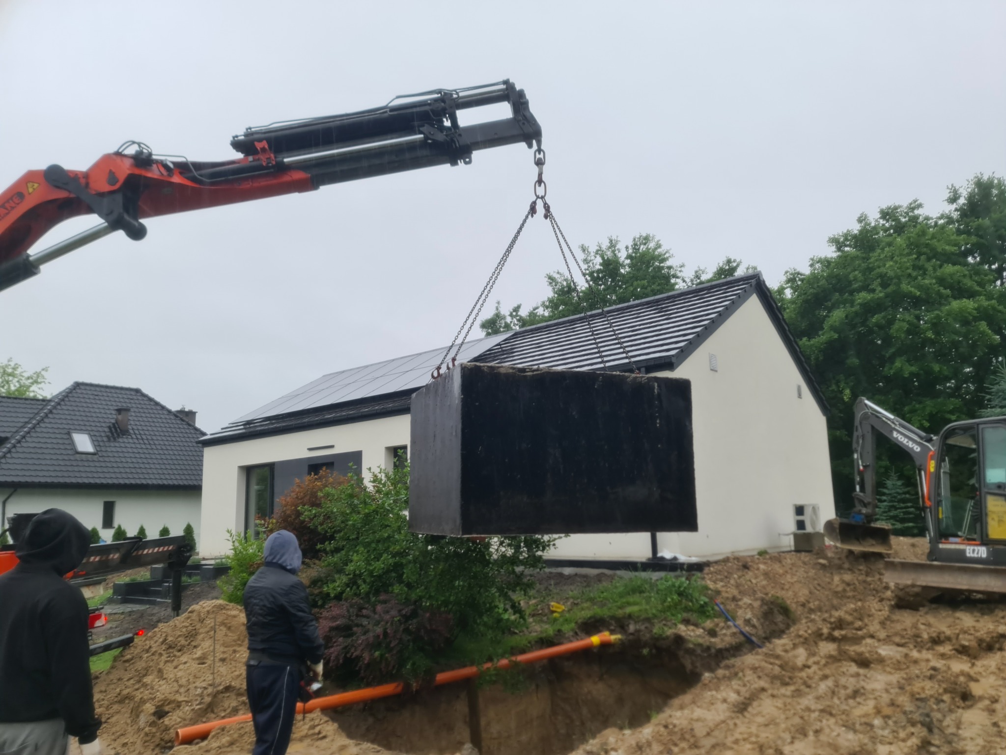 Ciężki, czarny zbiornik zawieszony na łańcuchach dźwigu, opuszczany do wykopu obok domu z panelami słonecznymi na dachu, w tle koparka Volvo, widoczni pracownicy w ubraniach roboczych.