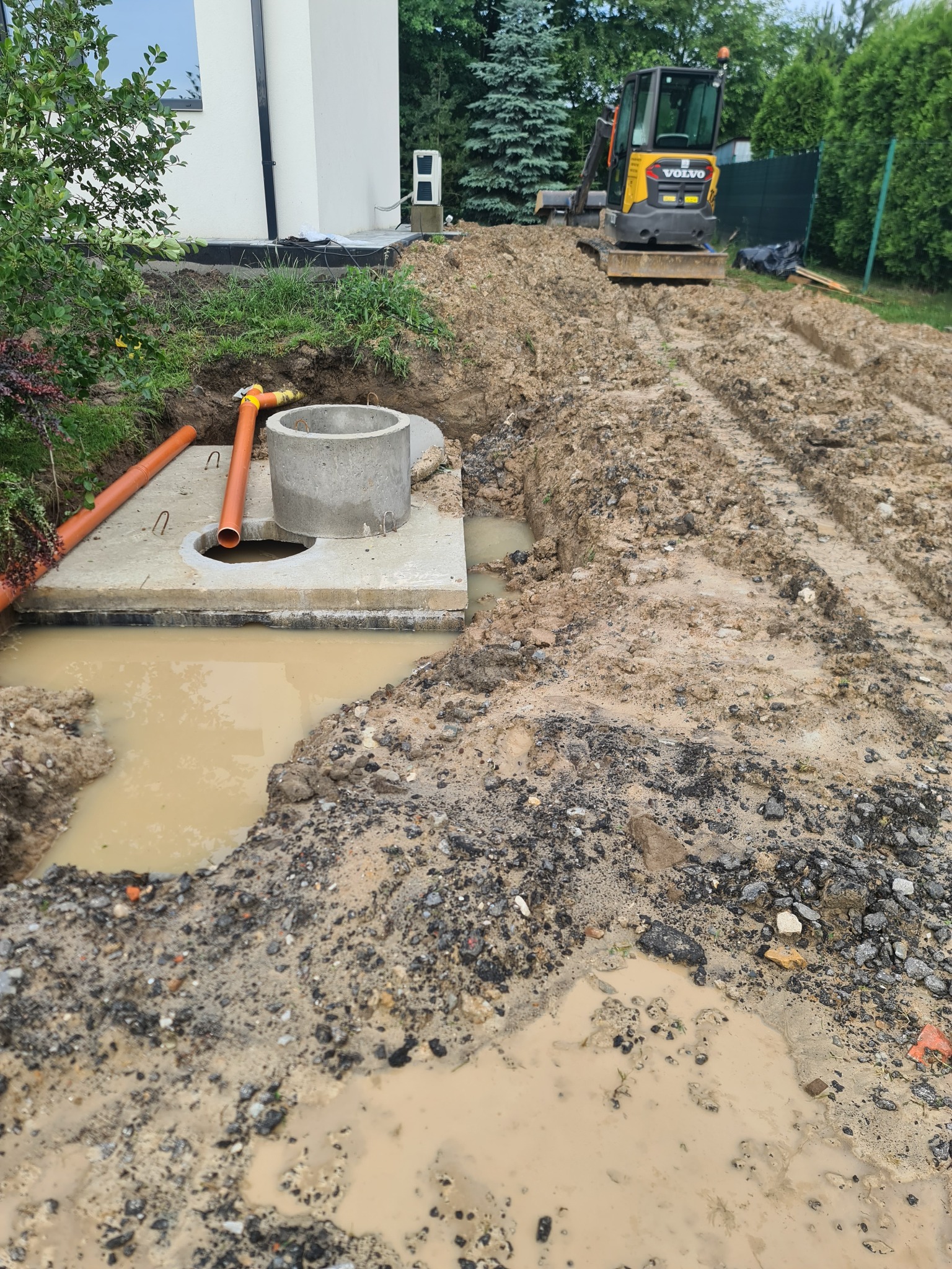 Wykonany wykop pod instalację z betonowym kręgiem i pomarańczowymi rurami kanalizacyjnymi, na ubitej ziemi widoczny mały żółty traktor.