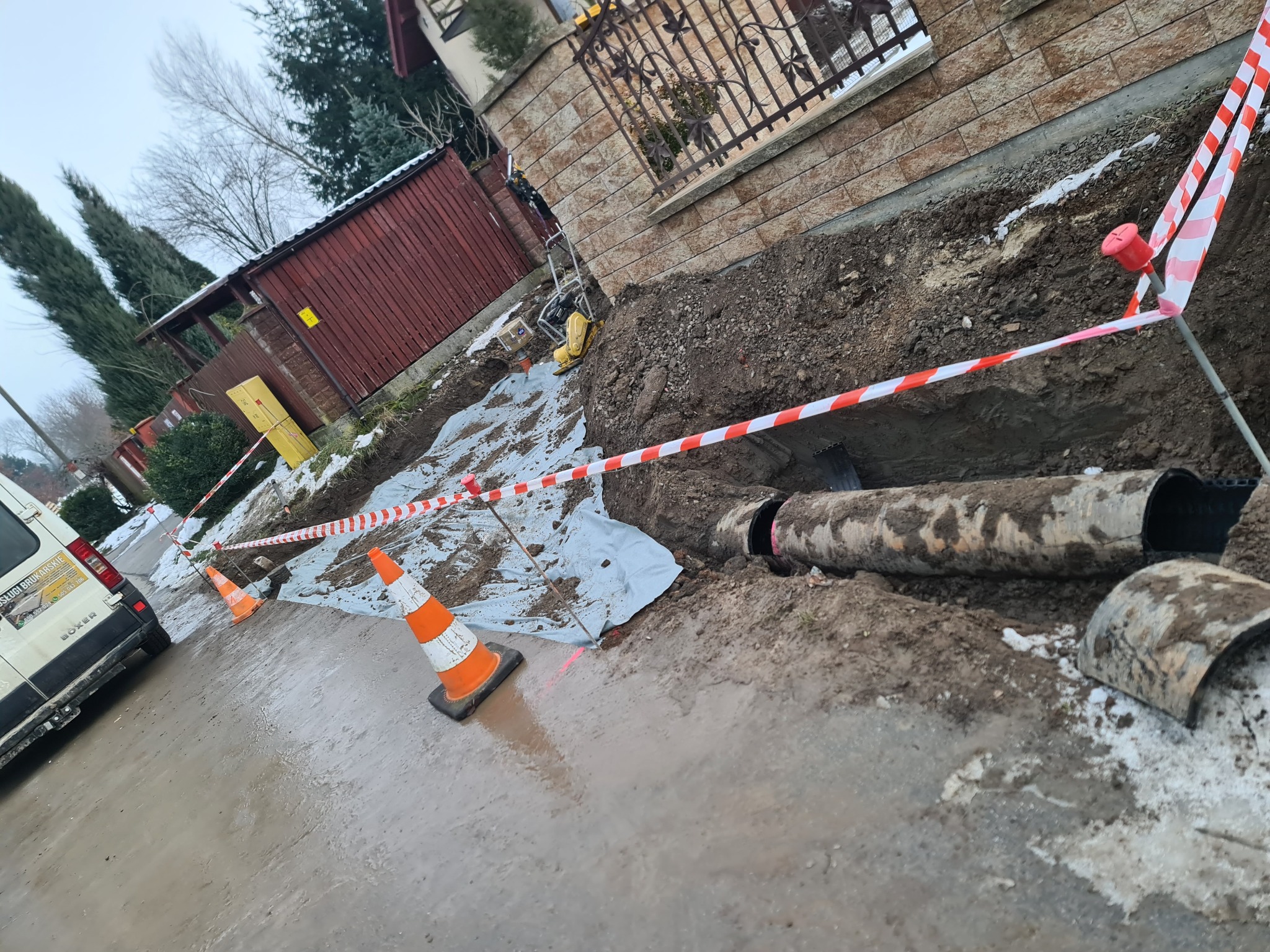 Odkryte rury kanalizacyjne w wykopie, zabezpieczone taśmą ostrzegawczą i pachołkami, częściowo przykryte folią, widoczny samochód dostawczy z reklamą usług brukarskich w tle.