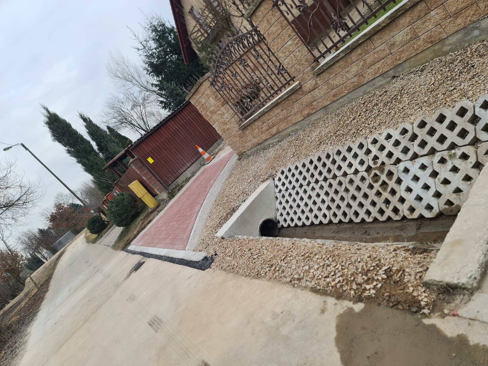 Ukończona ścieżka z czerwonej kostki brukowej z białym obramowaniem, obok ułożone ażurowe płyty betonowe, widoczny rów melioracyjny z kamieniami i wlotem kanalizacyjnym, tło: budynek z cegły...