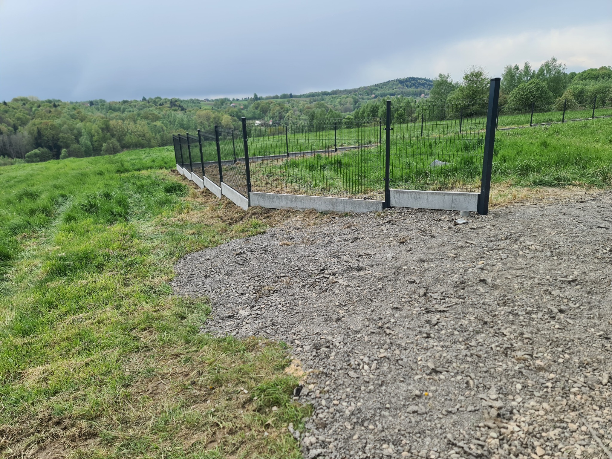 Nowo postawione ogrodzenie panelowe z podmurówką na nierównym terenie, widok z drogi szutrowej w kierunku zalesionych wzgórz pod pochmurnym niebem.