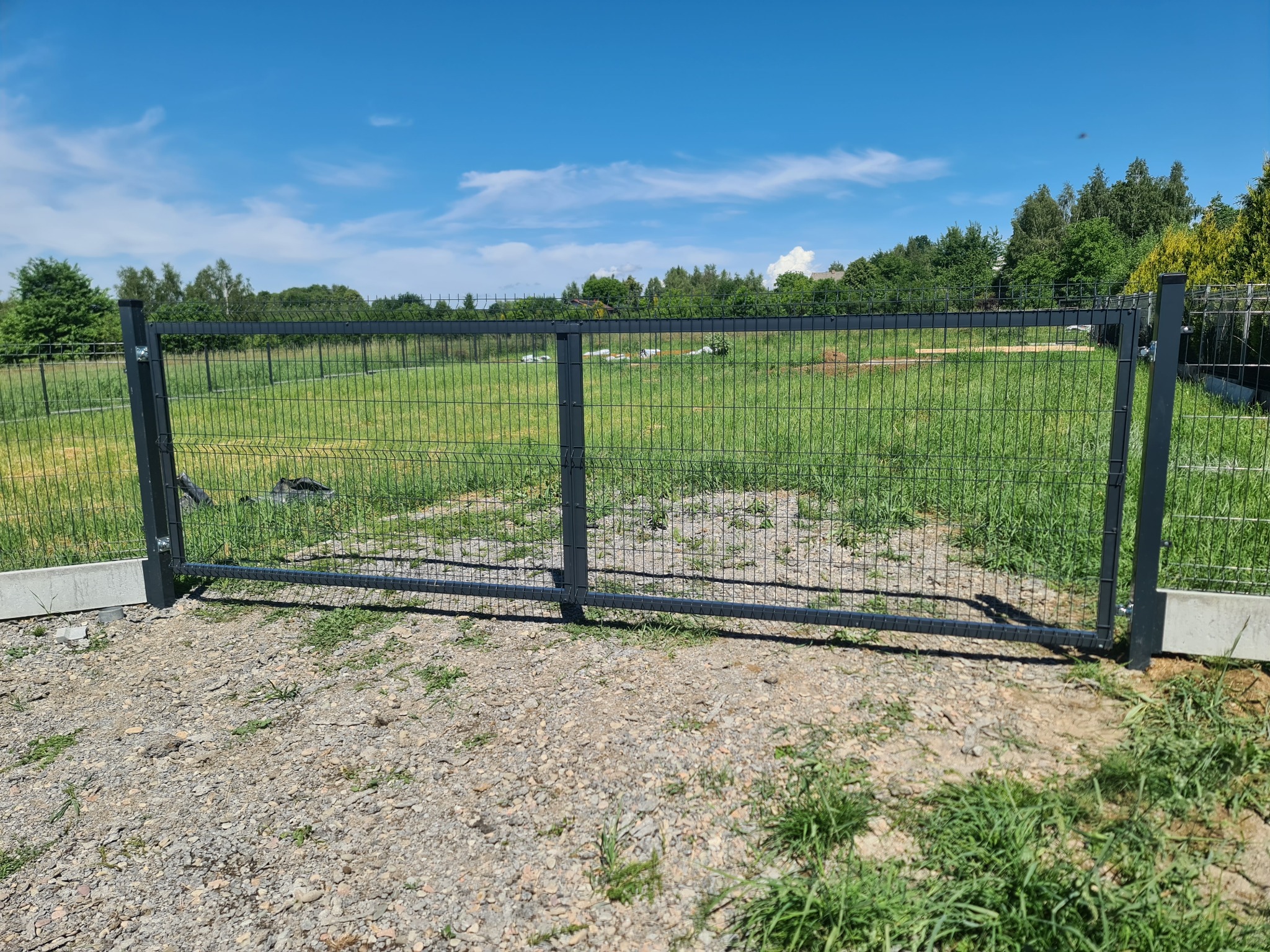 Nowoczesna, grafitowa brama dwuskrzydłowa z siatki panelowej na tle zielonego terenu i błękitnego nieba z delikatnymi obłokami.