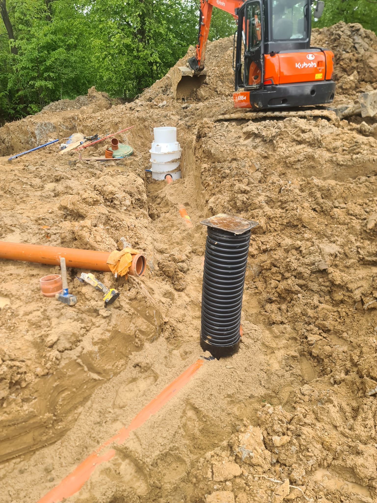 Wykonany wykop z ułożonymi rurami kanalizacyjnymi, studzienką rewizyjną i zbiornikiem, z pomarańczową koparką Kubota w tle, na terenie budowy.