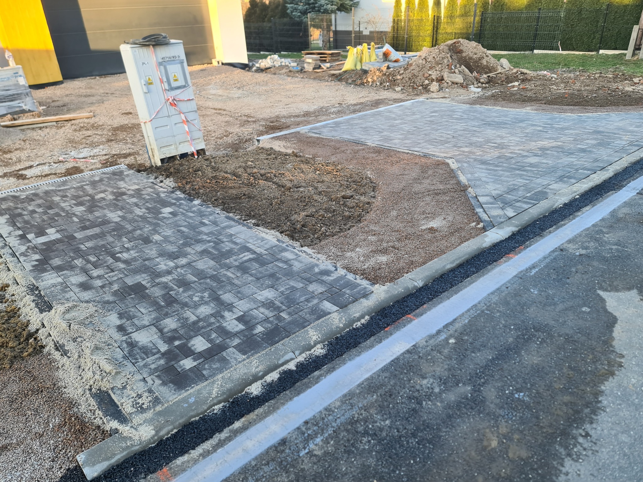 Fragment układanej nawierzchni z szarej kostki brukowej, widoczny piasek i podsypka, z boku skrzynka elektryczna zabezpieczona taśmą, w tle hałda ziemi i częściowo ukończony budynek.