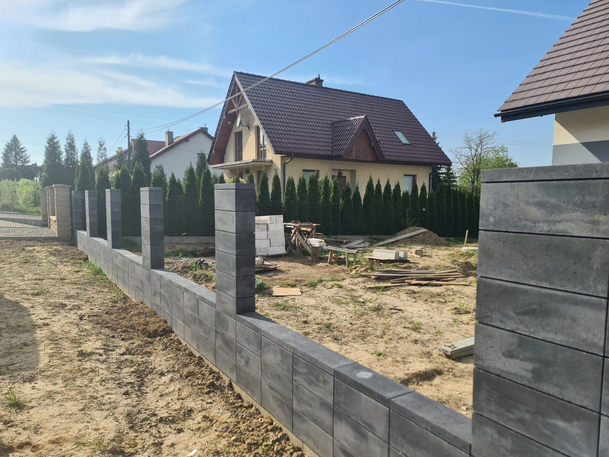 Wznoszony modułowy mur ogrodzeniowy z szarej kostki betonowej na tle domu jednorodzinnego i błękitnego nieba.