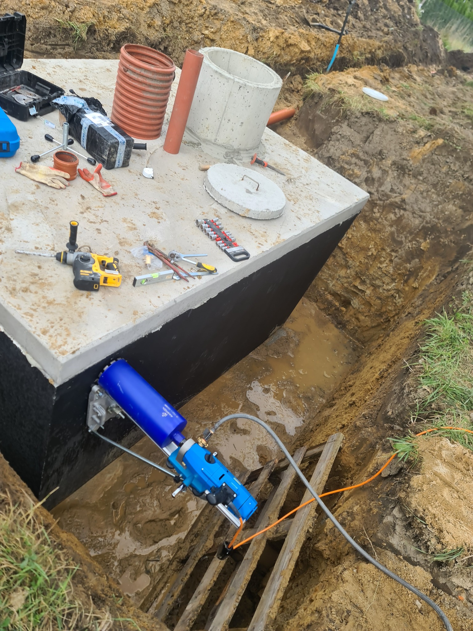 Betonowy zbiornik w wykopie, w trakcie wiercenia otworu wiertnicą diamentową, widoczne narzędzia i elementy instalacji.