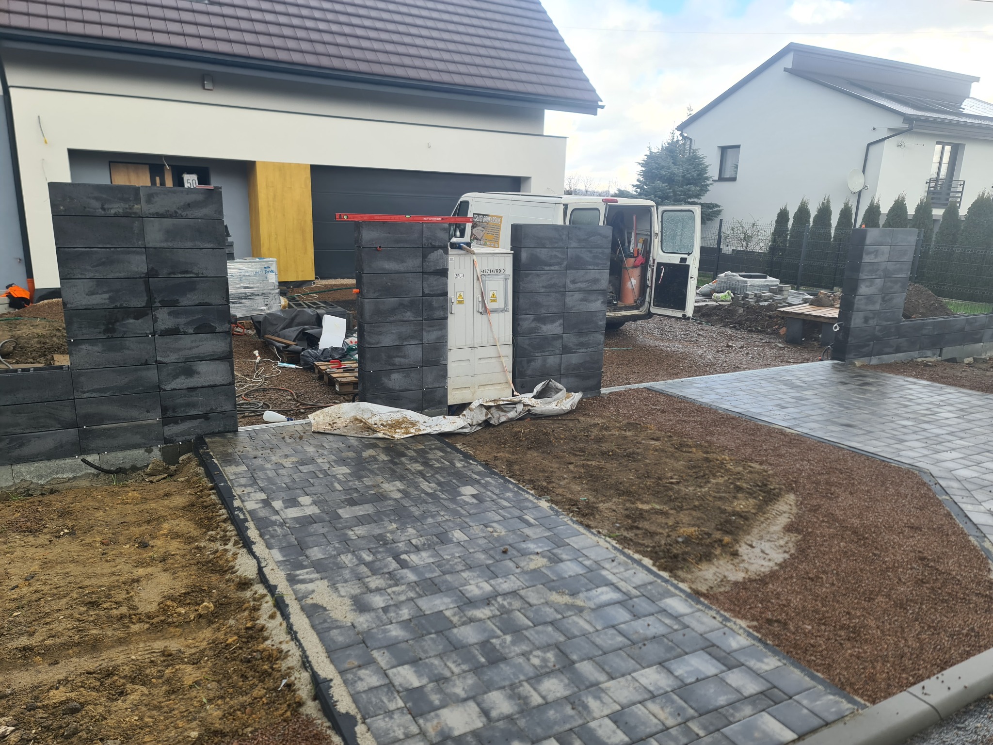 Budowa ogrodzenia z bloczków betonowych i układanie kostki brukowej na podjeździe do domu jednorodzinnego w trakcie realizacji, widoczny samochód dostawczy i skrzynka elektryczna.