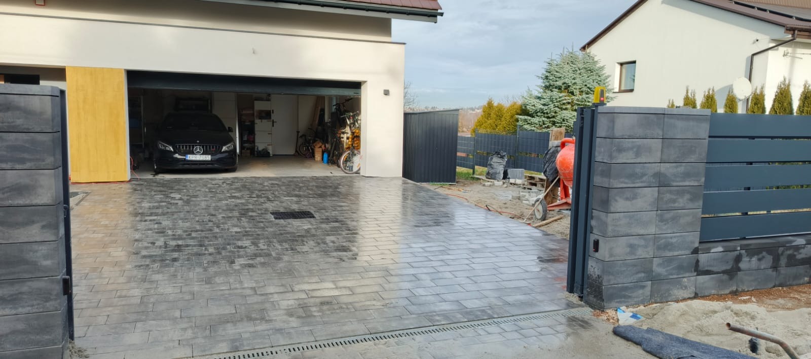 Nowoczesny podjazd z szarej kostki brukowej prowadzący do garażu z zaparkowanym czarnym Mercedesem, obok ogrodzenie panelowe z betonowymi słupkami, widoczna betoniarka i narzędzia.