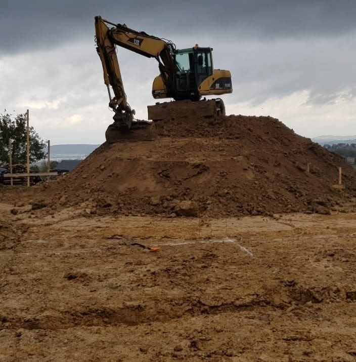 Żółta koparka CAT na szczycie pryzmy ziemi, widoczne drewniane paliki wyznaczające obszar robót, pochmurne niebo w tle.