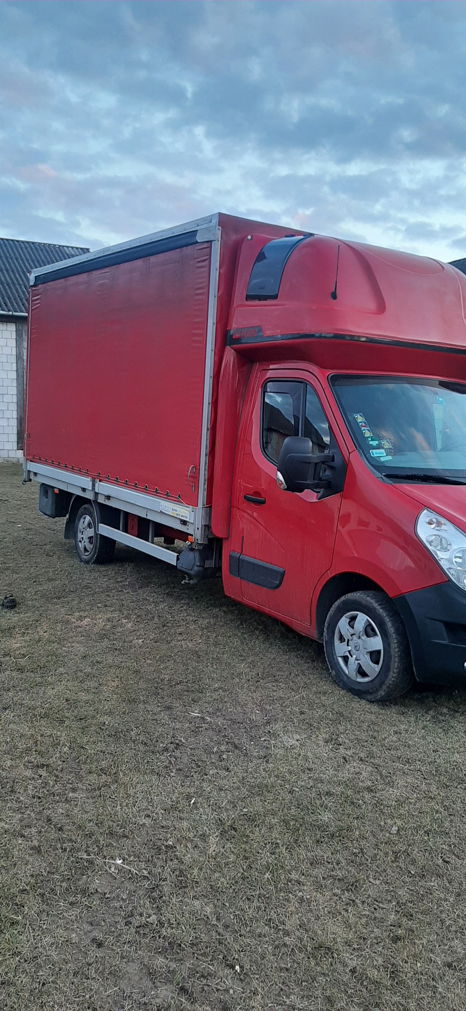 Czerwony samochód dostawczy z plandeką na trawiastym podłożu, widok z boku, pochmurne niebo w tle.