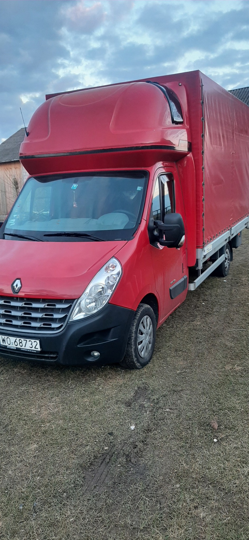 Czerwony samochód dostawczy Renault Master z plandeką i nadbudówką sypialną, zaparkowany na trawiastym podłożu, widoczne ślady opon.