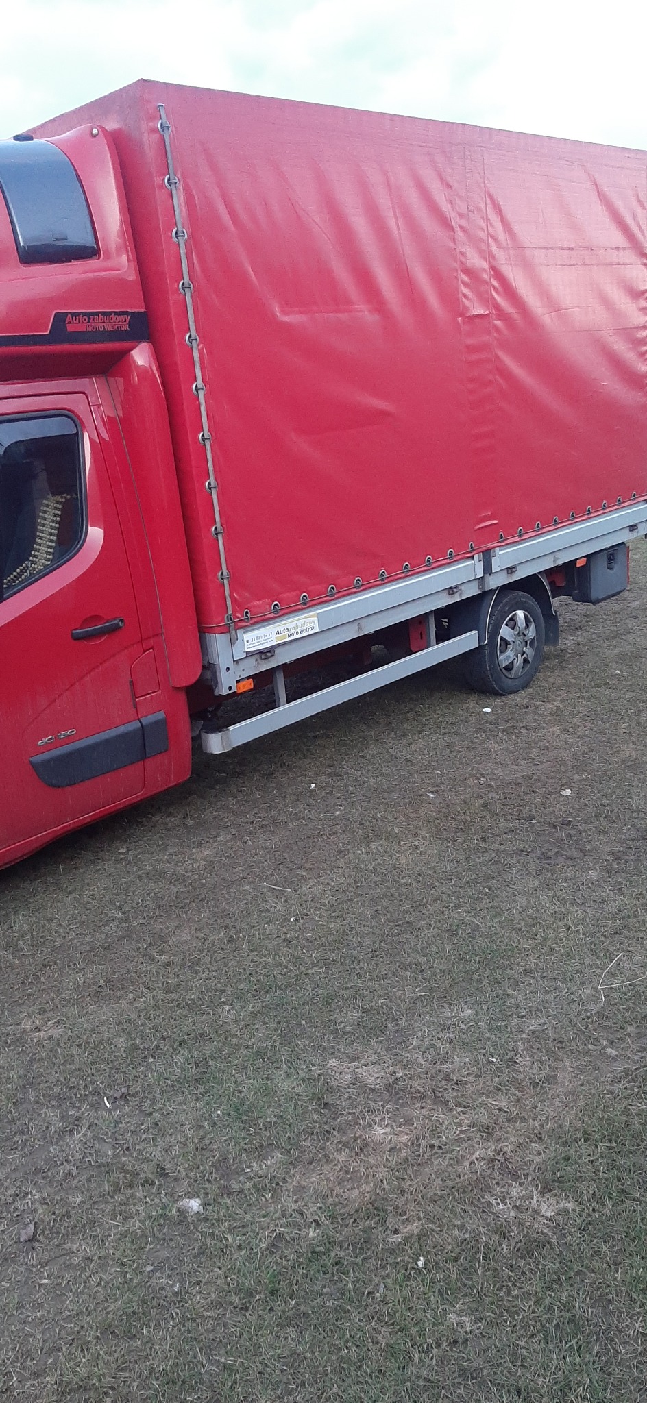 Czerwony samochód dostawczy z plandeką zaparkowany na trawiastym terenie, widoczny z boku, z logo 'Auto zabudowy MOTO WEKTOR' na kabinie.