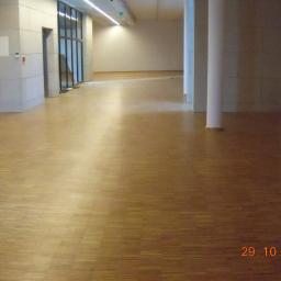 Ecologi Investment - Wnętrze komercyjne z parkietem ułożonym w jodełkę, jasne ściany z kolumnami i widoczne drzwi oraz okna w oddali.